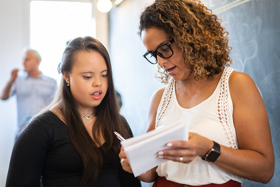 A student with Down syndrome and a woman wearing glasses review papers together in a classroom setting, with other people visible in the background near a chalkboard.